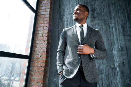 Handsome smiling african american manager in a suit buttons his jacket and looks to the side while standing against the background of the windowの写真素材