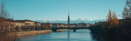 A picturesque landscape showcases a tranquil river flowing beneath a bridge, flanked by urban buildings and distant snow-capped mountains under a clear blue sky.の素材