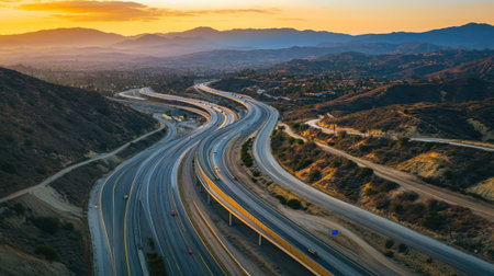 Aerial view of a dramatic mountain highway featuring multiple switchbacks, surrounded by rugged hills. The golden hour casts a warm glow over the landscape, enhancing its natural beauty.の素材