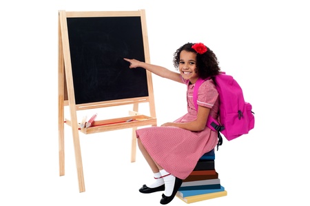 Pretty school girl sitting on stack of books and pointing towards blank chalkboardの写真素材