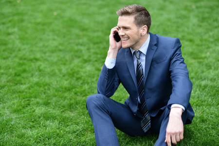 Businessman sitting on meadow talking on smart phoneの写真素材