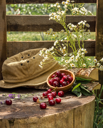 Cherries in a bowl lie on a stump next to a hat and flowersの写真素材