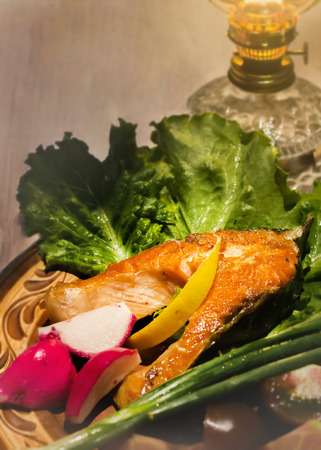 One fried salmon steak lies on the leaves of the salad on the background of a kerosene lamp. The radish and green onionsの写真素材