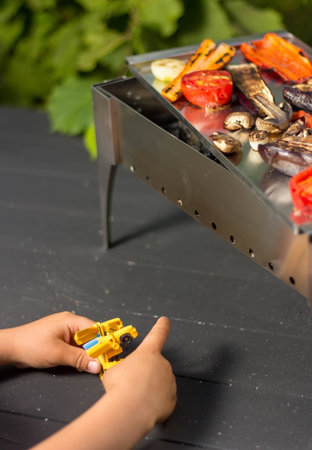 Grilled vegetables on the barbecue, next to the child plays a robot. A child and a robot are waiting for dinner to be preparedの写真素材
