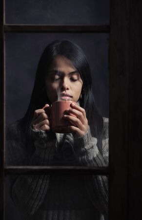 Young woman behind window with a cup of coffee or tea, focus on handsの写真素材