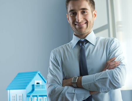 Young real estate agent with arms crossed and model house on background.の写真素材