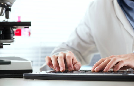 Researcher working at desk and typing on a keyboard with microscope and laboratory glassware.の写真素材