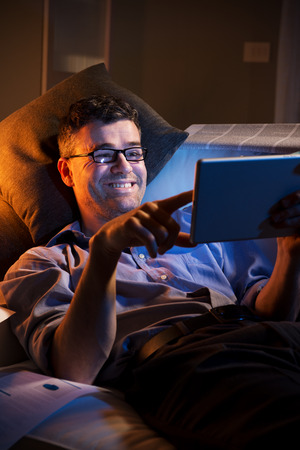 Man working at night lying down on sofa in the living room with tablet.の写真素材