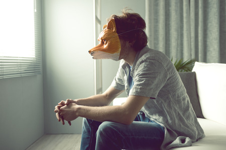 Young man wearing a fox mask sitting on sofa in front of a window.の写真素材