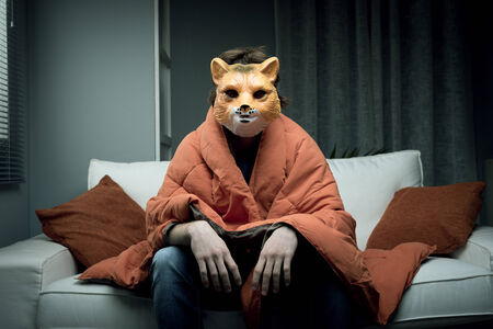 Young man wearing a fox mask sitting on sofa covered with a blanket.の写真素材