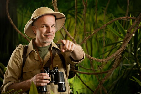Smiling explorer in the jungle with binoculars pointing at camera.