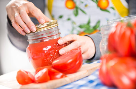 Woman filling glass jars with tomato sauce on checked tablecloth, hands close-up.の写真素材