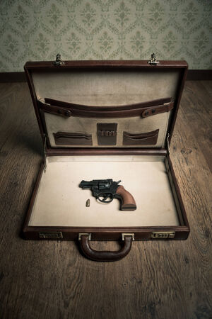 Open vintage leather briefcase with only revolver gun and bullet inside, vintage wallpaper and wooden floor on background.の写真素材