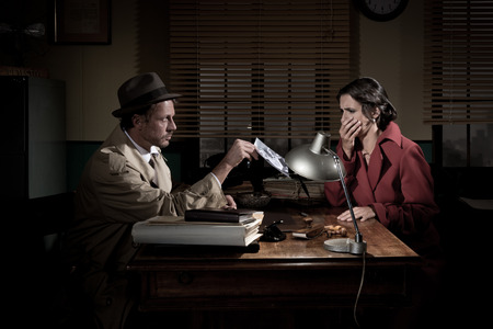 Handsome detective at office desk showing a picture to a young woman, film noir scene.の写真素材