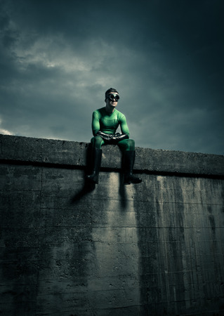 Green pensive superhero sitting on a concrete wall with dramatic cloudy sky on background.の写真素材