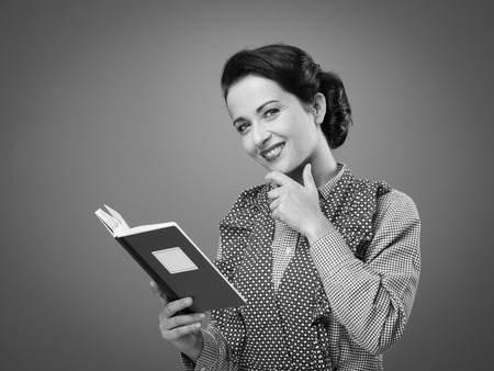 Smiling retro woman in apron with cookbook and hand on chin