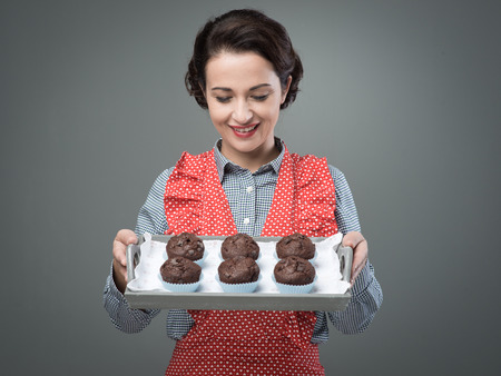 Smiling vintage woman in apron serving homemade chocolate muffins on a trayの写真素材