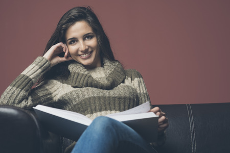 Young relaxed woman on sofa reading a book and smilingの写真素材