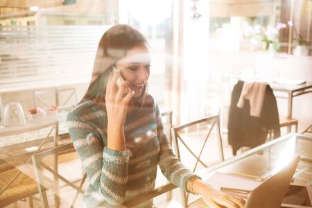 Young woman having a phone call and working at computer next to a windowの写真素材