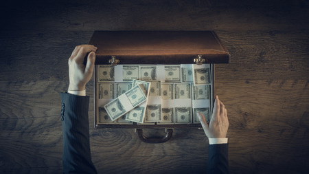 Rich businessman opening a leather briefcase filled with dollar packs, top view, unrecognizable...