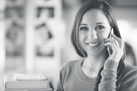 Cute young student girl having a phone call with books on backgroundの写真素材