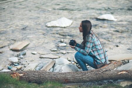 Young tourist woman exploring nature and holding a digital camera sitting along the riverの写真素材
