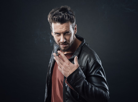 Confident fashionable man in leather jacket smoking a cigarの写真素材