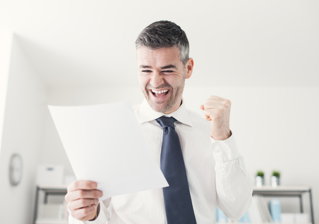 Cheerful businessman in the office receiving good news, he is holding a contract document and raising his fistの写真素材