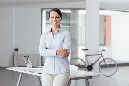Beautiful woman leaning on office desk and smiling at cameraの写真素材
