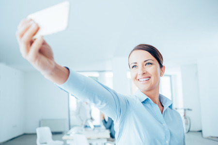 Smiling business woman taking a selfie in her office using a smart phoneの写真素材