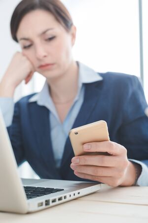 Pensive businesswoman working at office desk and reading sms on her mobile phone