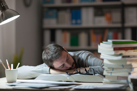 Young man sleeping at his desk late at night, he is leaning on a book and holding glasses, stress and exhaustion conceptの写真素材