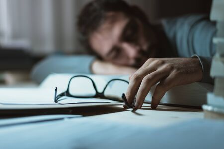 Young man sleeping at his desk late at night, he is leaning on a book and holding glasses, stress...