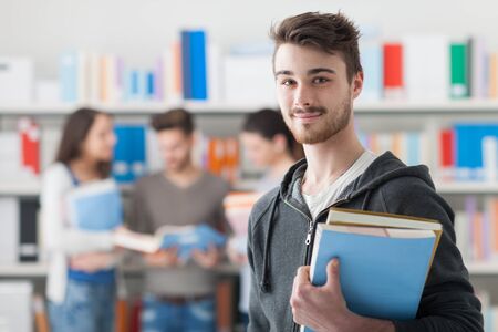 Confident handsome student holding books and smiling at camera, library bookshelves on background, learning and education conceptの写真素材