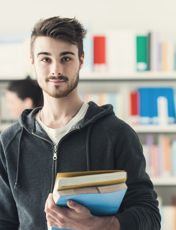 Confident handsome student holding books and smiling at camera, library bookshelves on background, learning and education conceptの写真素材