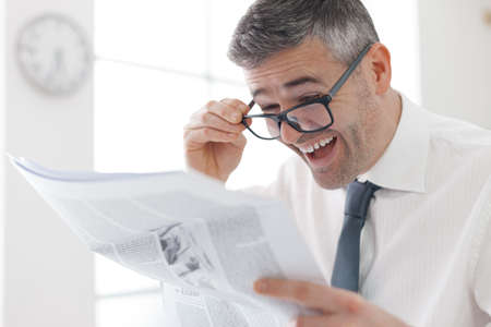 Businessman in the office, reading bad breaking news on a financial newspaper, he is shocked and adjusting his glassesの写真素材