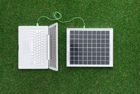 Laptop on the grass connected to a solar panel, alternative energy sources and electrical power generation conceptの写真素材