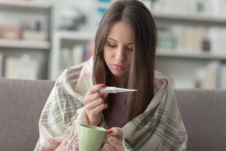 Sick young woman at home on the sofa, she is covering with a blanket, holding an hot drink and...