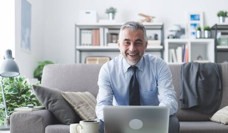 Businessman working at home, he is sitting on the couch, working with his laptop and smiling at cameraの写真素材