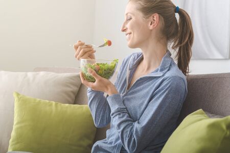 Young blonde woman relaxing on the couch at home and eating a fresh garden salad, healthy lifestyle and nutrition conceptの写真素材