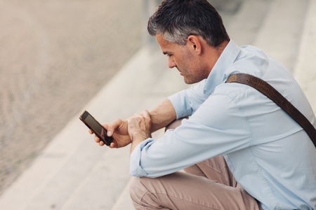 Man in the street, he is sitting on a step and using a smart phoneの写真素材