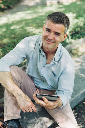 Confident man at the park connecting with a digital tablet, he is sitting on a bench and smiling at cameraの写真素材