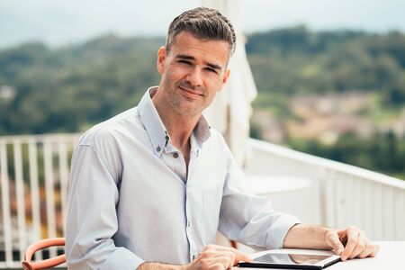 Man relaxing on the terrace and using a digital touch screen tablet, panorama view on the backgroundの写真素材