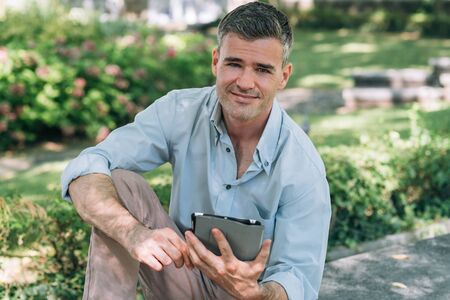 Confident man at the park connecting with a digital tablet, he is sitting on a bench and smiling at cameraの写真素材