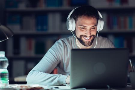 Young smiling man networking and connecting to internet using a laptop late at night, he is sitting...