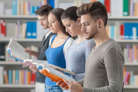 Group of college students in the library standing in line, holding books and reading, bookshelves on backgroundの写真素材