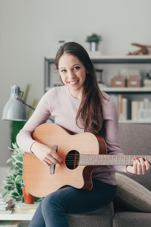 Smiling beautiful girl sitting on the sofa in the living room and playing guitar, music and leisure conceptの写真素材