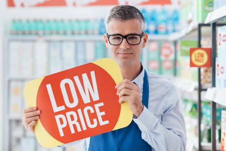 Confident supermarket clerk holding an advertisement discount sign, store shelves on the backgroundの写真素材