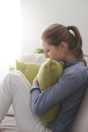 Lonely depressed woman at home, she is sitting on the sofa and hugging a pillowの写真素材