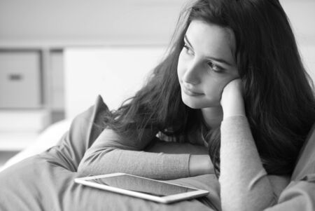 Smiling young woman relaxing on the bed at home, she is using a tablet and lying downの写真素材